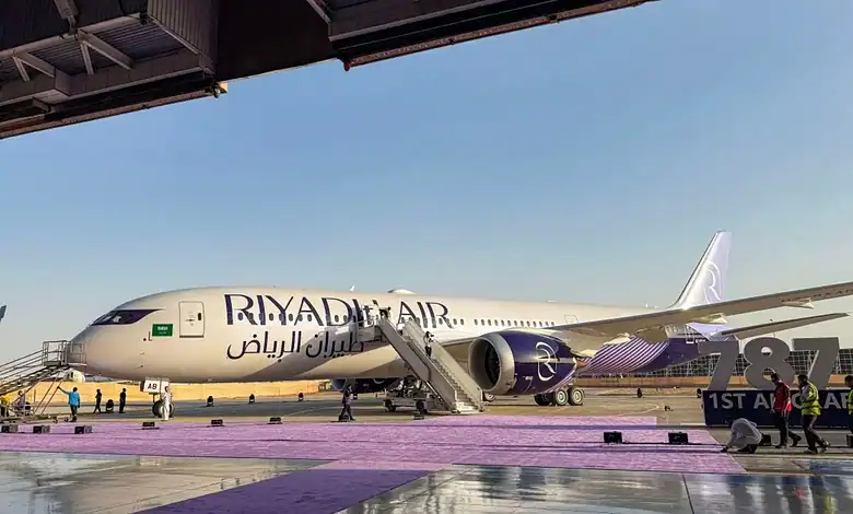 Riyadh Air aircraft parked at the airport with stairs and ground crew nearby.