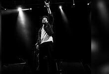 Samay Raina performing on stage with microphone, raising hand, in black and white.