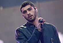 Zayn Malik singing on stage with a microphone, wearing a dark jacket and looking into the distance.